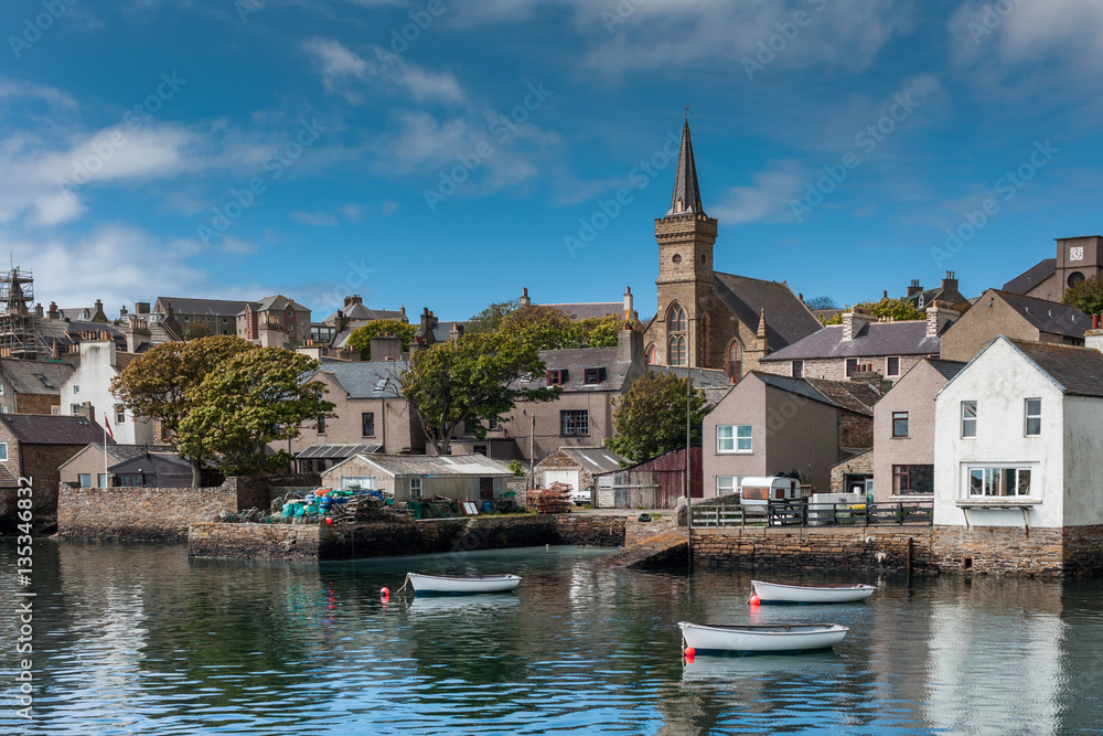 Fototapeta premium Orkneys, Scotland - June 5, 2012: View on the old cluttered docks with parish church towering over gray and brown houses under blue sky. Sloops reflecting on sea water. Green vegetation.