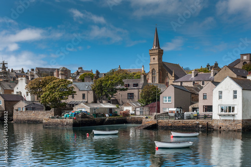 Orkneys, Scotland - June 5, 2012: View on the old cluttered docks with parish church towering over gray and brown houses under blue sky. Sloops reflecting on sea water. Green vegetation. photo