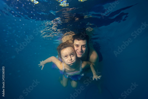 The daughter and mother play sports and swim underwater on a blue background, looking at the camera and smiling. Portrait. The view from under the water. Horizontal view