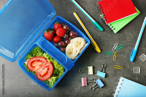 Tasty sandwich and fruits in lunchbox and stationery on dark background