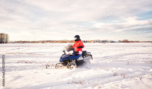 A teenage girl in bright colored winter gear and a helmet snowmobiling across a snow covered field with a farmyard in the background in a winter landscape