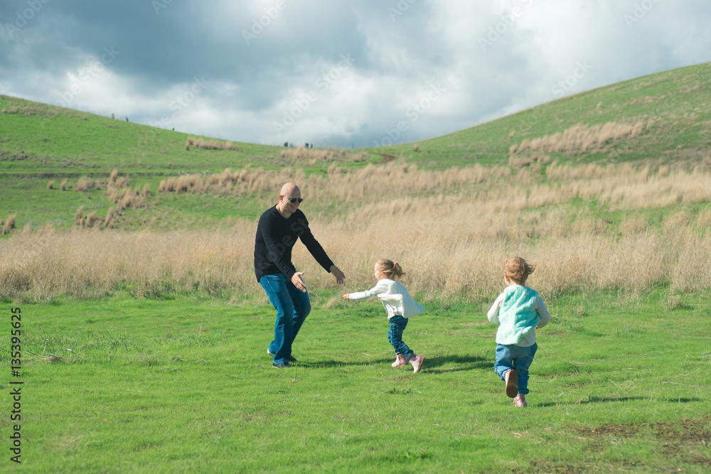 Smiling father playing and running with his two laughing daughters in the park on green grass. Beautiful view with stormy clouds over their heads.