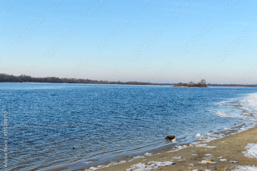 Winter landscape. A view of the wide river. On an empty shore - chunks of ice. Cold season. Clear day.