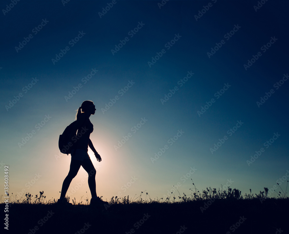 silhouette Young woman tourist with backpack goes on the background on sunset sky