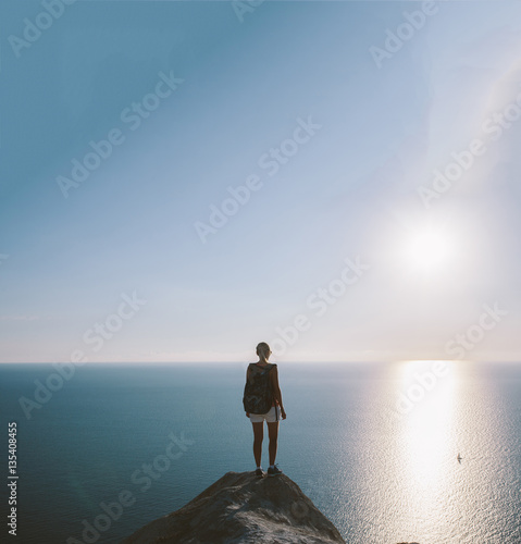 Young woman hiker with backpack standing on cliff and looking forward on the background of the sea, sky. lady tourist on top of a mountain enjoying view...