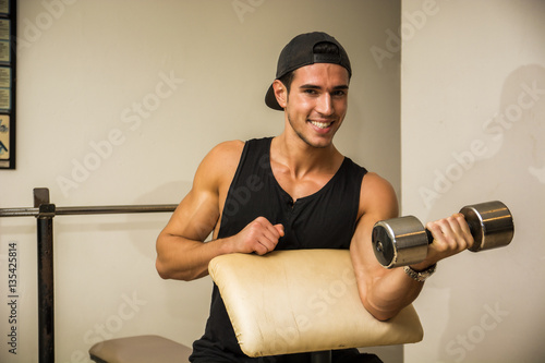 Handsome muscular young man exercising biceps in gym with dumbbells, smiling and looking at camera