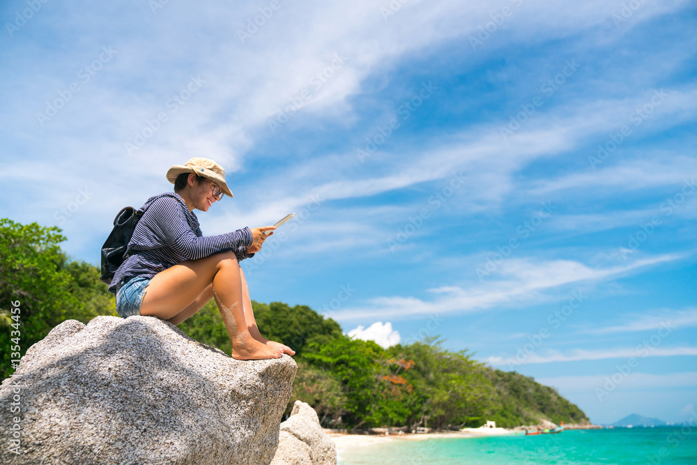 hiker girl in a hat with a backpack with glasses sitting on a rock on the island of sandy beach with a clipboard in his hands.