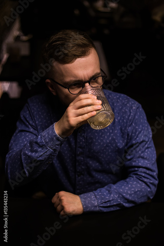 Business man, in blue shirt and glasses, drinking cocktail of vintage old fashion glass in loft bar