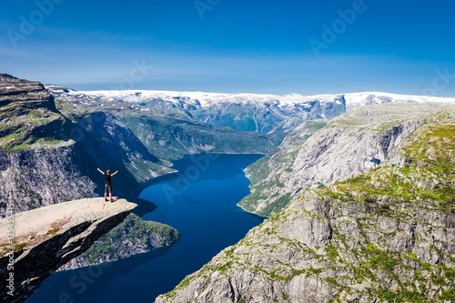 Panoramic view of standing man on Trolltunga cliff, Norway, Scandinavia. Sunny day with amazing blue sky. Majestic mountains in the background. Clear blue water. 