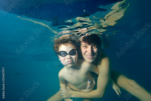 The child and coach swim underwater in the pool on a blue background. Look at the camera and smiling. The view from under the water. Close-up. Landscape orientation