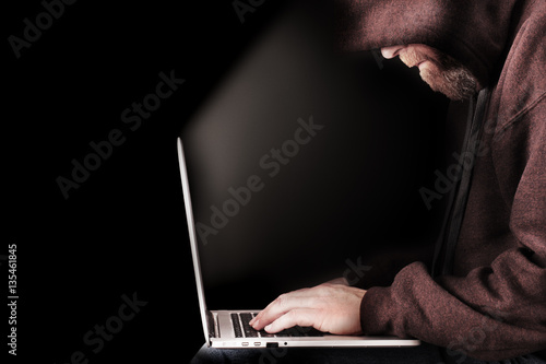Male computer hacker wearing a hooded top leaning over a laptop in the dark. The screen light illuminates the man with a beard performing illegal activities. Black copy space on the left