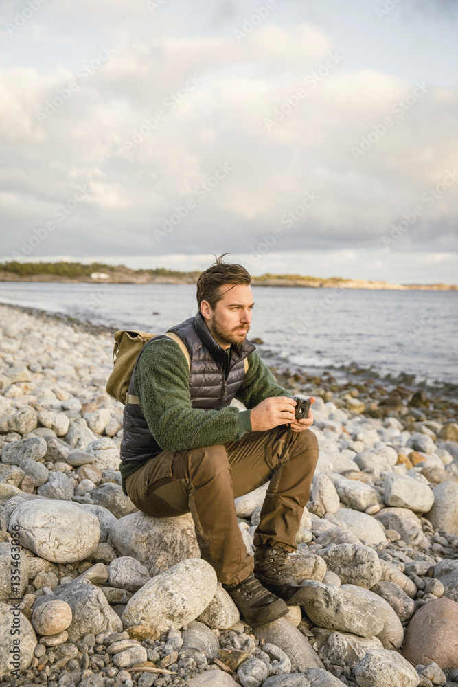 Man sitting on rock with camera at beach against sky Man sitting on rock with camera at beach against sky