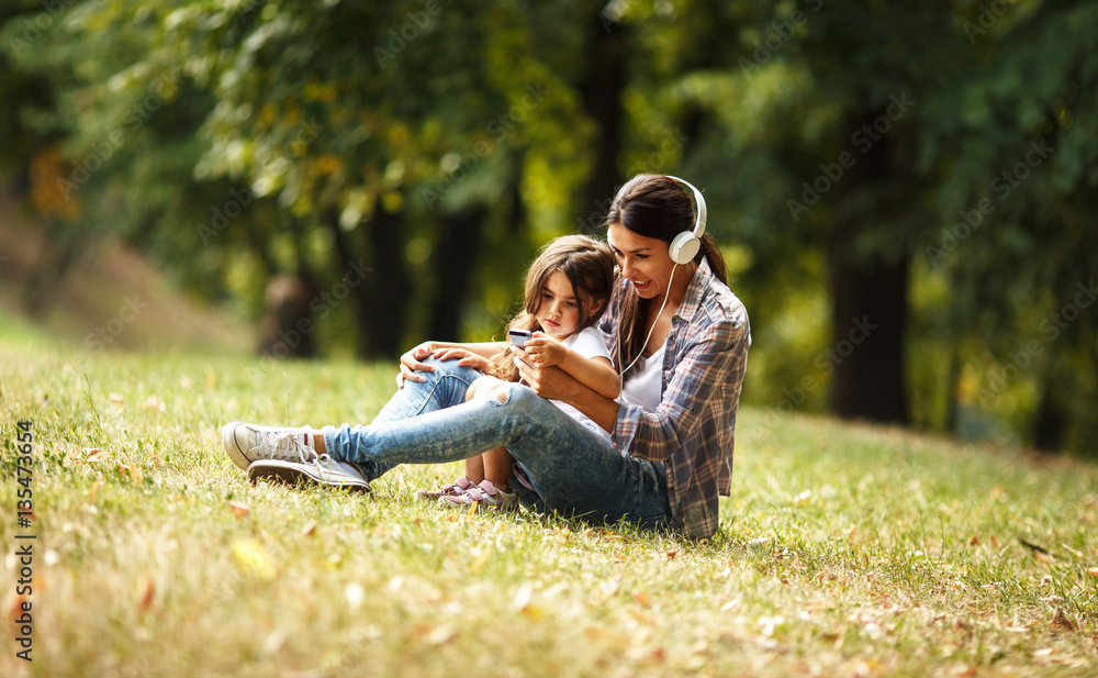 © BalanceFormCreative - Mother and daughter sitting on grass and listening to music on smart phone. © BalanceFormCreative - Mother and daughter sitting on grass and listening to music on smart phone.
