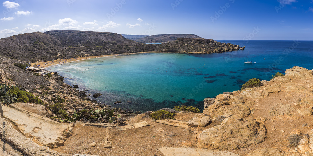 Malta - Panoramic view of Ghajn Tuffieha sandy beach with sail boat, blue sky and crystal clear green sea water 