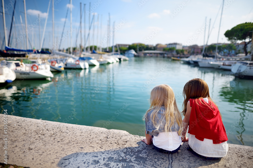 Two cute little sisters watching small yachts in harbor in Desenzano del Garda on beautiful autumn day