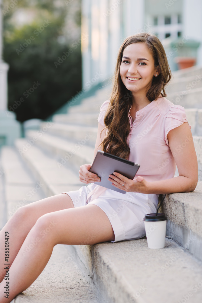 Obraz premium Attractive young woman sitting on some steps with her tablet and coffee to go