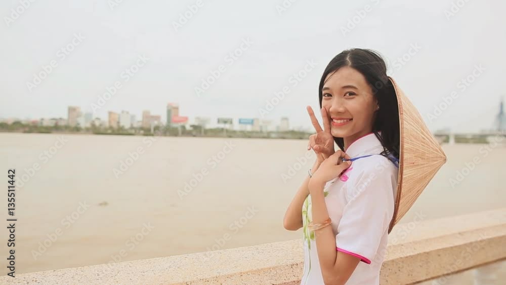 Vietnam girl in the national costume and dress Ao Dai posing on the waterfront of the city of Da Nang. On the back of the cap Non La. Vietnam girl in the national costume and dress Ao Dai posing on the waterfront of the city of Da Nang. On the back of the cap Non La.