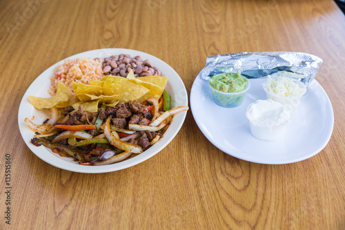 Steak fajitas with peppers and onions with a side of chips, pinto beans, rice, foil wrapped tortillas, sour cream, guacamole, and cheese on white plate on wooden table