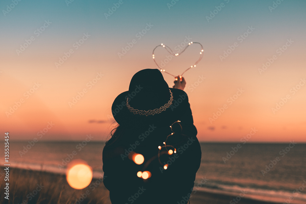 Cuore luminoso tenuto in mano da una ragazza vestita di nera sulla spiaggia  al tramonto. San Valentino Concetto. Stock Photo