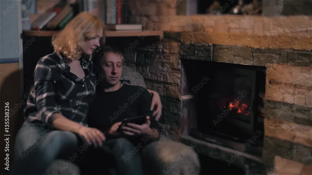 Young Couple Sitting On Sofa Near Fireplace At Winter Season At Home, Man Using Tablet PC Young Couple Sitting On Sofa Near Fireplace At Winter Season At Home, Man Using Tablet PC