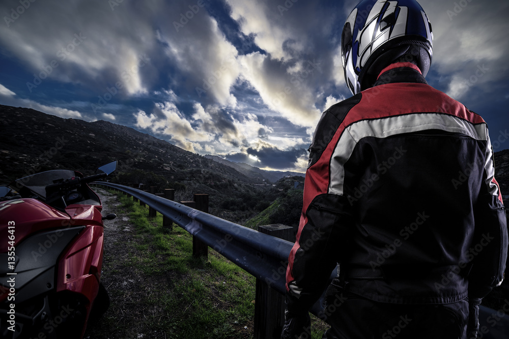 Fototapeta premium Male motorcyclist wearing protective leather racing suit with a red bike or motorcycle on an open road. The vehicle is cropped to become generic non branded. The image depicts travel and adventure.