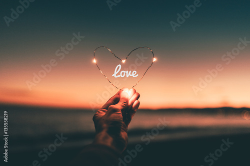Fotografija Simbolo del cuore illuminato con scritta love per San Valentino tenuto in mano di fronte al tramonto sul mare