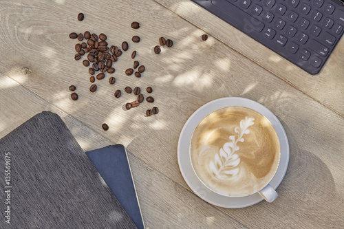 Cup of coffee latte with coffee beans with electronic device and notebook on light wooden background. Top view.