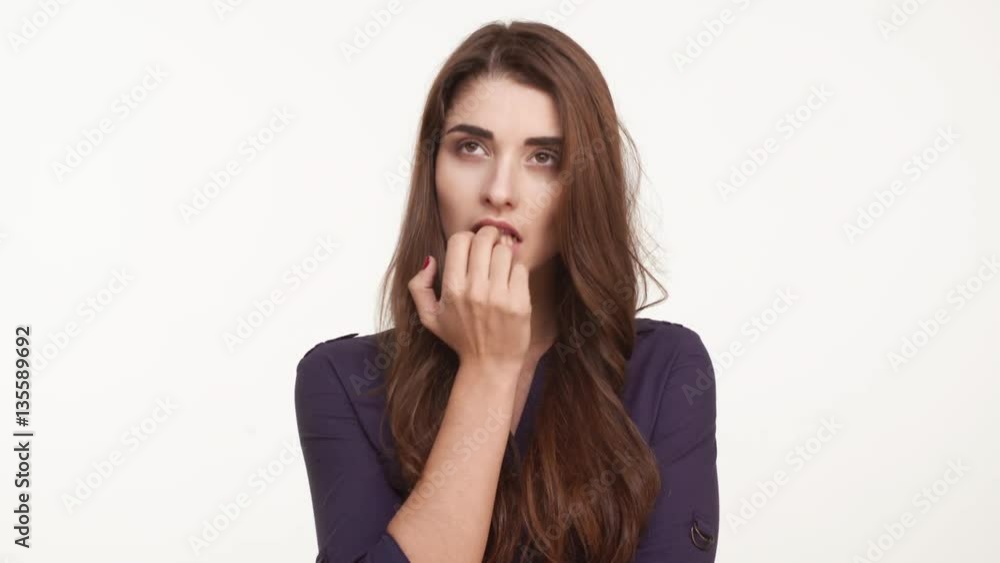 Nervous beautiful Caucasian female with long brown hair wearing dark blue shirt standing kicking her heels rubing hands biting nails on white background Nervous beautiful Caucasian female with long brown hair wearing dark blue shirt standing kicking her heels rubing hands biting nails on white background