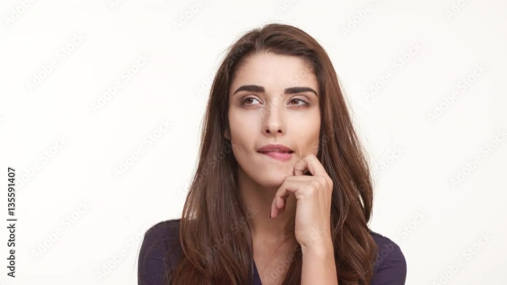 Thoughtful attractive young Caucasian female in dark blue shirt with long brown hairstanding on white background biting lip touching hair then coming to mind Thoughtful attractive young Caucasian female in dark blue shirt with long brown hairstanding on white background biting lip touching hair then coming to mind