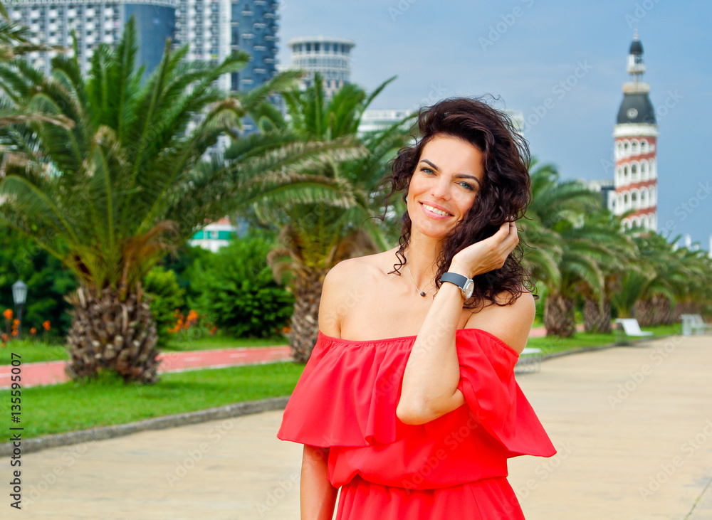 Fototapeta premium Woman corrects hair and smile in the long red dress. Pretty girl surrounded by tropical palms near sea