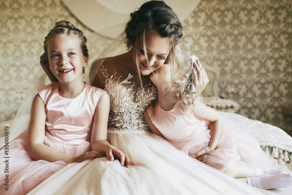 Young bride in ivory dress sits with little girl on bed