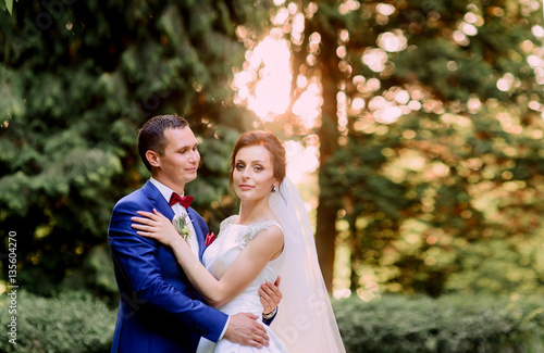 Warm rays of evening sun shine behind happy wedding couple huggi