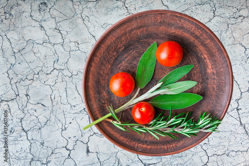 Sage and rosemary green leaves and cherry tomatoes on ceramic plate on marble surface. Top view.