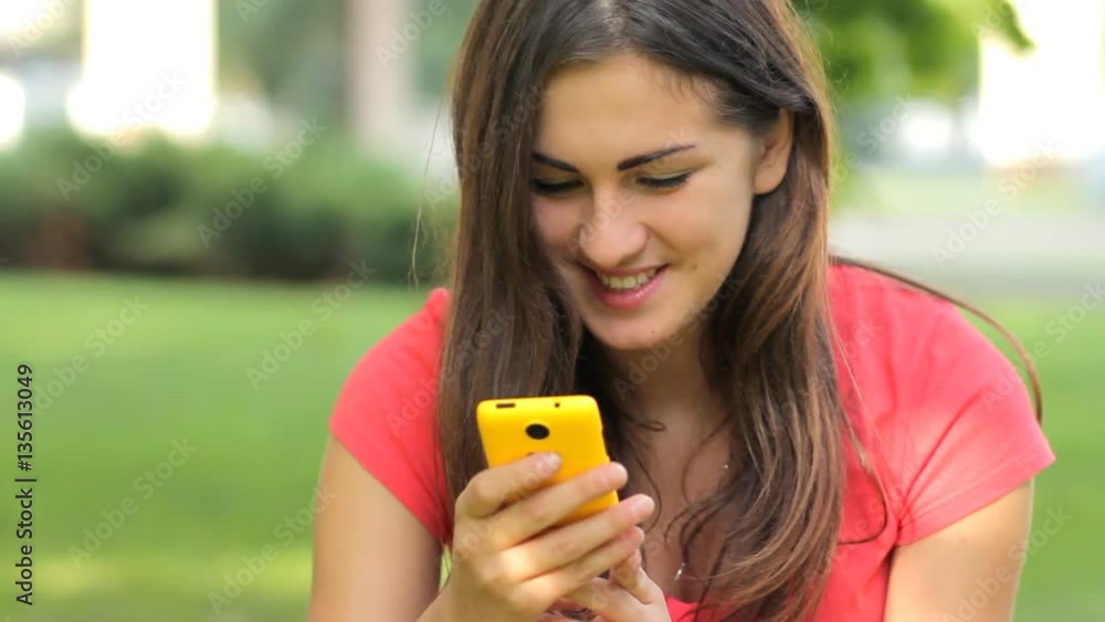 Smiling attractive young woman sms texting using app on smart phone in park Smiling attractive young woman sms texting using app on smart phone in park