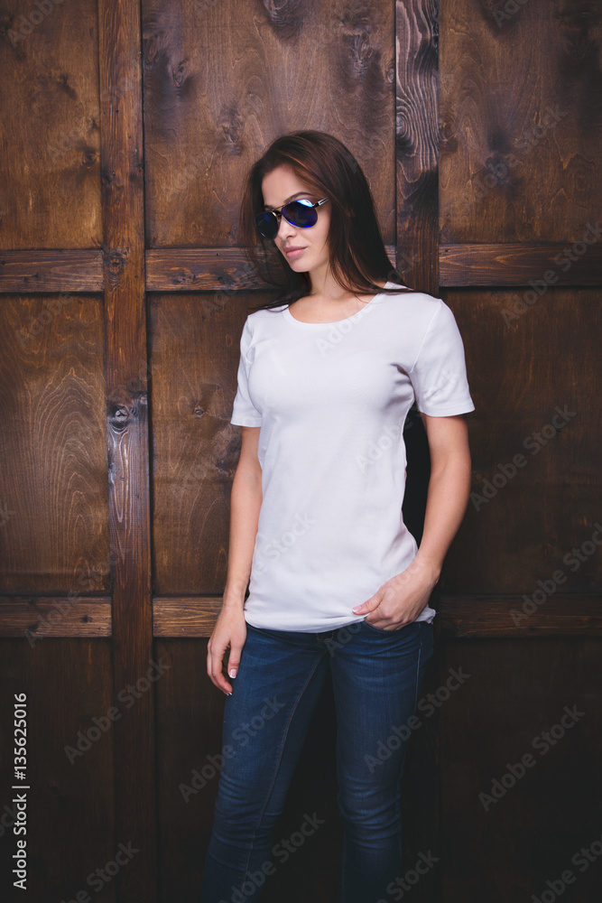 Fototapeta premium Young woman wearing blank white t-shirt with area for your logo or design, mock-up of template white t-shirt, wooden wall in the background