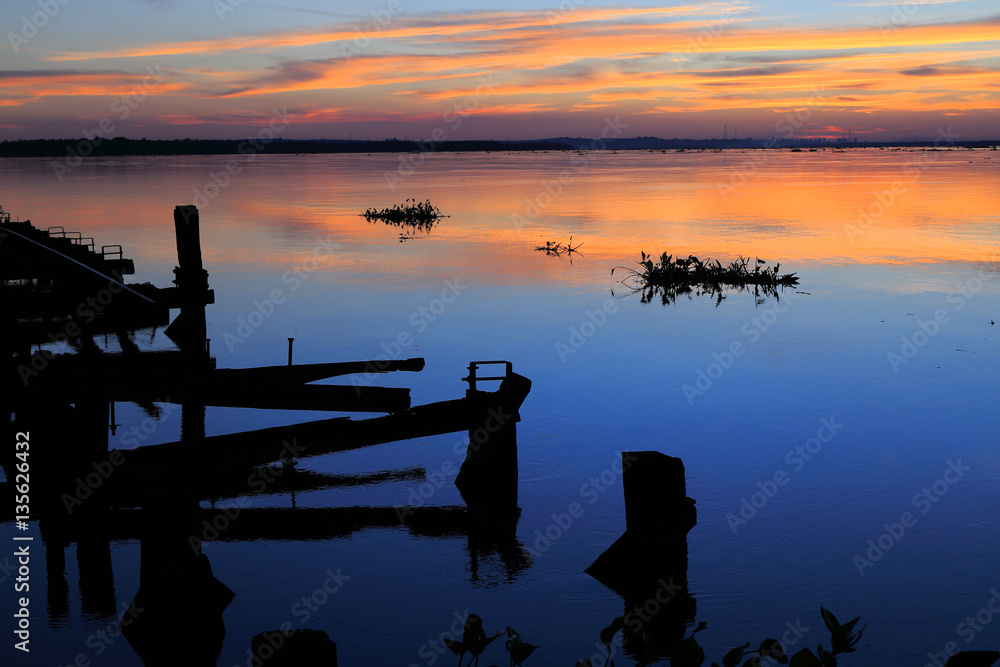 Beautiful sunset on the river parana in entre rios, Argentina, south america. An old wooden pier, the sky in orange tones is reflected on the water and aquatic plants pass by the river. - obrazy, fototapety, plakaty 