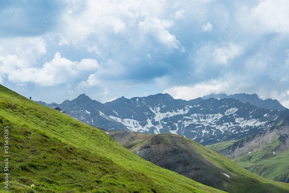 Cloudy sky over green hills in the Italian Alps. A tiny hiker can be seen on the hill. Cloudy sky over green hills in the Italian Alps. A tiny hiker can be seen on the hill.