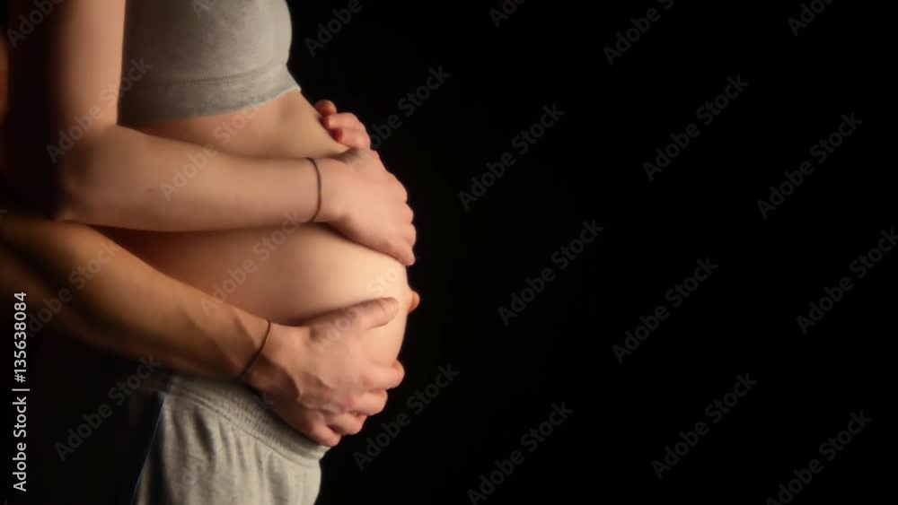 Close-up shot of pregnant woman smoothing the bare belly and future father holding gently his wifes hands. Touching moments of pregnancy black background Close-up shot of pregnant woman smoothing the bare belly and future father holding gently his wifes hands. Touching moments of pregnancy black background