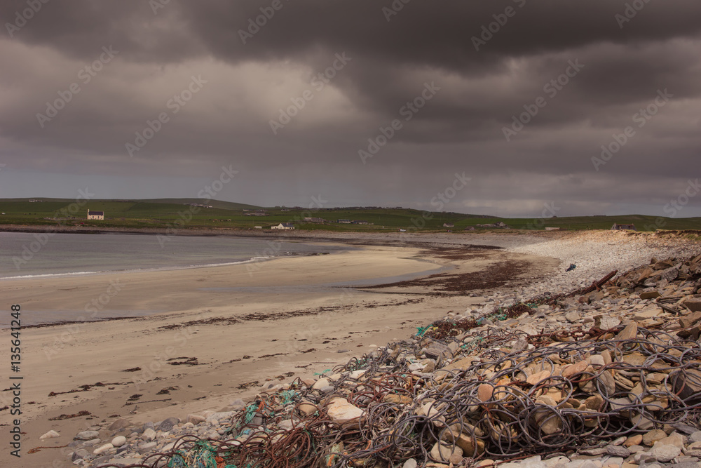 Orkneys, Scotland - June 5, 2012: The beach at Skara Brae Neolithic Settlement under heavy dark stormy sky. Sand, rocks and rubble. Quiet ocean and a green band of meadows on the horizon.