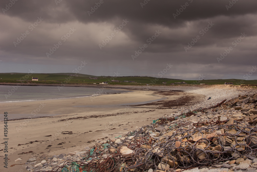 Orkneys, Scotland - June 5, 2012: The beach at Skara Brae Neolithic Settlement under heavy dark stormy sky. Sand, rocks and rubble. Quiet ocean and a green band of meadows on the horizon. Orkneys, Scotland - June 5, 2012: The beach at Skara Brae Neolithic Settlement under heavy dark stormy sky. Sand, rocks and rubble. Quiet ocean and a green band of meadows on the horizon.