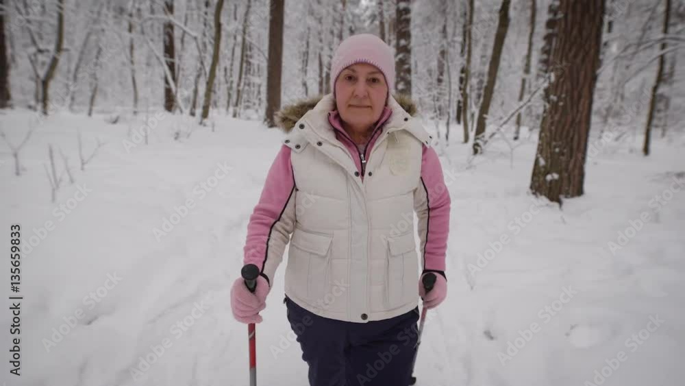 Nice grandma walks on the snowy forest, where there are trees, pines and other . She energetically moving forward along the path. Woman dressed in warm sportswear.