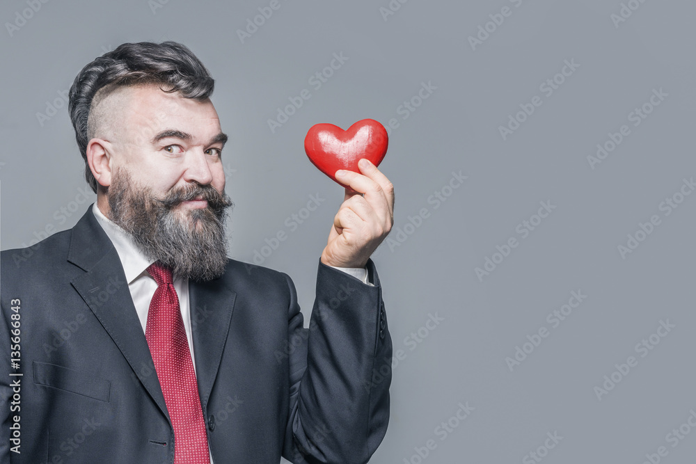 Smiling adult bearded man in the suit and red tie holding a red heart. Gray background Smiling adult bearded man in the suit and red tie holding a red heart. Gray background