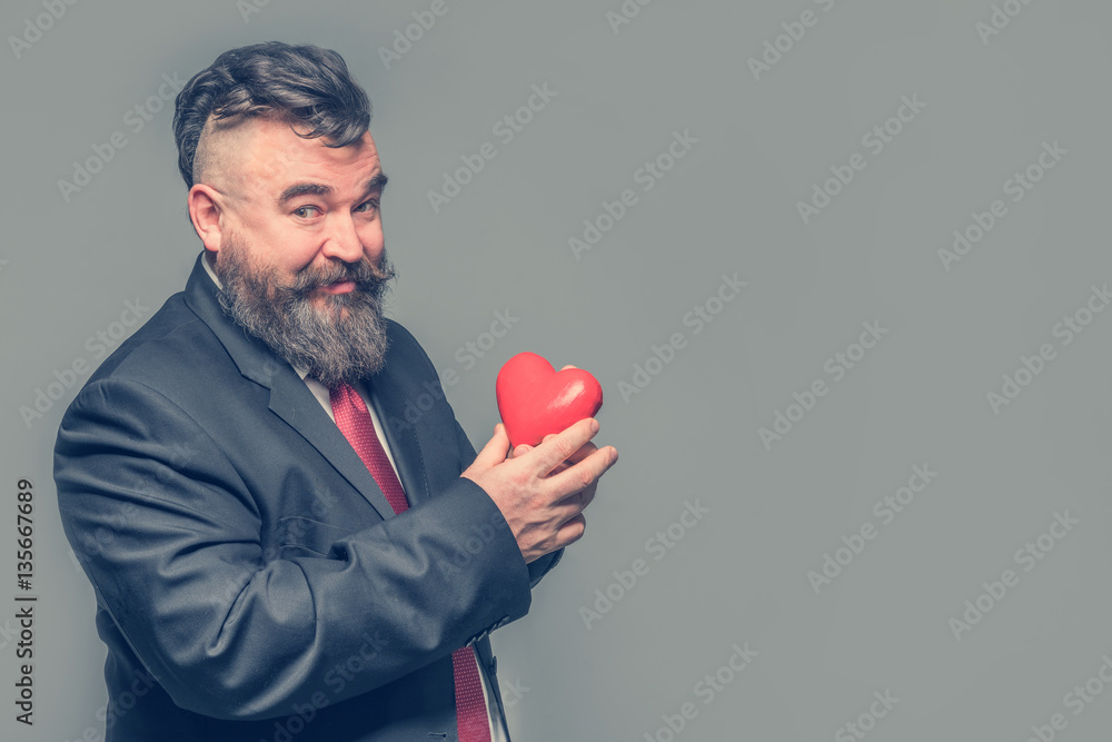 Smiling adult bearded man in a suit holding a red heart on a gray background. Isolated Smiling adult bearded man in a suit holding a red heart on a gray background. Isolated