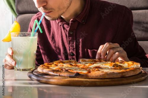 Having lunch and soft drink at pizzeria. Hungry male person drinking lemonade and taking slice of pizza at restaurant on bright sunny day