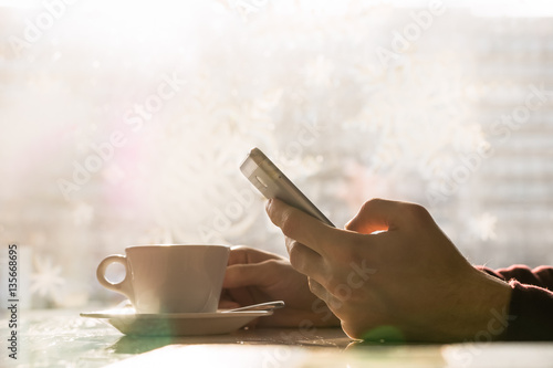 Using smartphone and having coffee at cafe. Close-up image of hands holding phone at dinner table with cup of coffee in front of window with bright sunlight