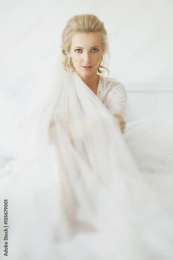 Bride in long white veil sits on large soft bed Bride in long white veil sits on large soft bed