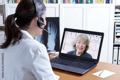 Telemedicine concept: female doctor in her surgery office with headset in front of her laptop, an x-ray of a foot in hand, talking with a senior patient.
