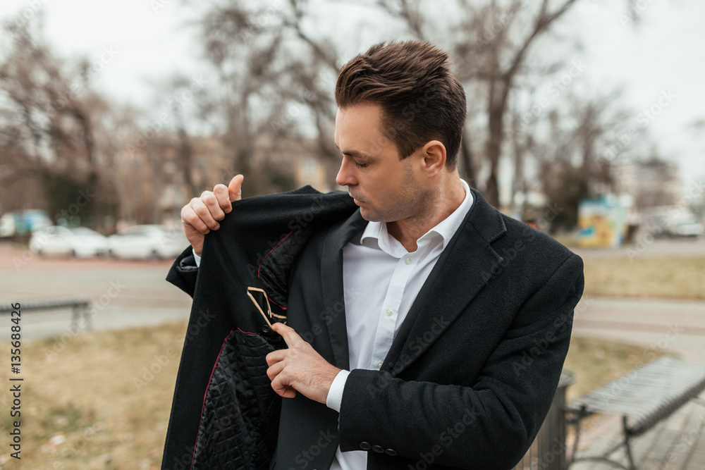 the man in a black coat and a suit costs on the street. He looks for  something in an inside pocket of a coat. He has very serious look. Stock  Photo