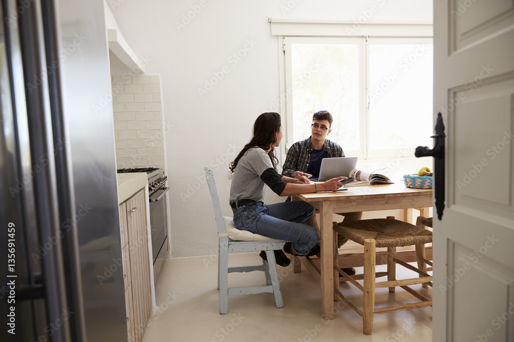 Two teenagers using laptop computer talking at kitchen table