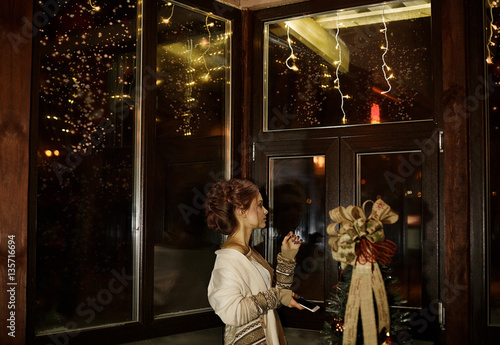 Beautiful girl with pretty hairstyle stands with phone in hands near the big wooden window in cozy, comfortable home, apartments. Cute girl look out window and wait for her husband, wait for call.